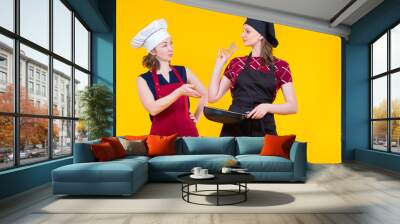 Women in chef's clothes. Girls discuss cooking. Cooking college student holding frying pan in her hand. Concept of training for restaurant chef. Two female chef's on yellow. Career restaurant workers Wall mural