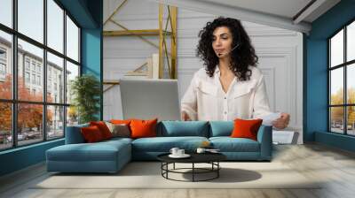 Smiling Indian employee with curly hair in headphones talks via video link, holds a web conference with colleagues, an ethnic worker in a headset works on a laptop at home Wall mural