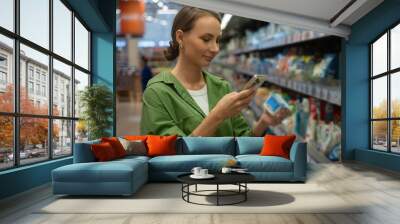 Young woman is comparing cleaning products in a supermarket. She is using a smartphone to scan barcodes and learn more about the products Wall mural