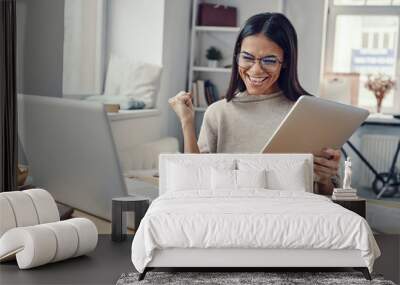 Beautiful young woman in casual clothing using laptop and cheering while working indoors Wall mural