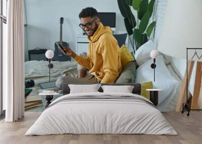 Cheerful African man using technologies while sitting on the bed at home Wall mural