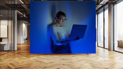 Confident young woman working on laptop against a wall with blue lighting Wall mural