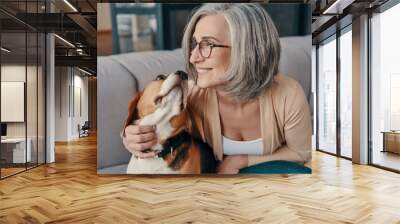 Happy senior woman in casual clothing spending time with her dog while sitting on the sofa at home Wall mural