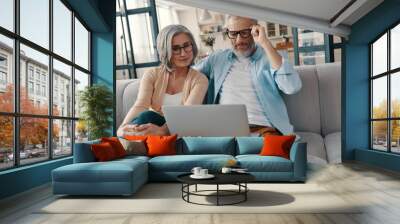 Modern senior couple in casual clothing smiling and using laptop while bonding together at home Wall mural