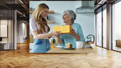 surprised senior woman receiving a gift box from her daughter while sitting at the kitchen together Wall mural