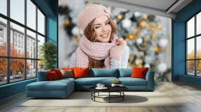 Beautiful young redhead woman with blue eyes and long curly hair,in a beige knitted hat,scarf and gloves posing in Studio on light background ornate Christmas tree with yellow balls Wall mural