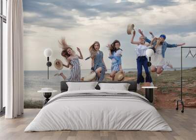 Five attractive and fun young girls having fun on the beach. Teenage sisters are resting together on the rocky shore of the blue ocean on a cloudy day against the blue sky and the coastline. Wall mural