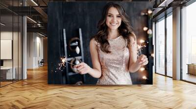 Girl with a sparkler near the Christmas tree. A cheerful young woman with a cute smile in a beige dress is standing and holding a sparkling sparkler in the hands against the background of the Christma Wall mural