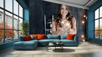 Girl with a sparkler near the Christmas tree. A cheerful young woman with a cute smile in a beige dress is standing and holding a sparkling sparkler in the hands against the background of the Christma Wall mural