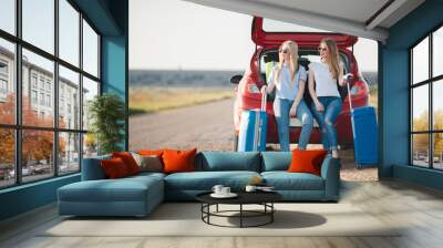 Young, slim, beautiful women, a blonde in a blue dress and a red-haired girl in a red dress,long black eyelashes, two best friends are preparing to travel on a red car with suitcases and things Wall mural