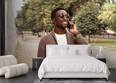 Portrait happy smiling african man calling on smartphone wearing brown knitted cardigan and sunglasses on city street over park background Wall mural