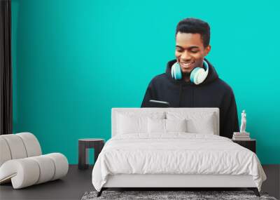 Portrait of happy smiling young african man with smartphone and headphones listening to music wearing black hoodie isolated on blue background, copy space Wall mural