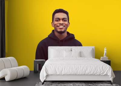 Portrait of modern smiling young african man looking at camera wearing black hoodie isolated on yellow background Wall mural