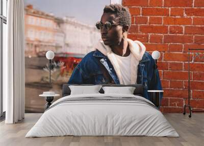 Portrait of stylish young african man model wearing denim jacket posing on a city street over brick wall background Wall mural