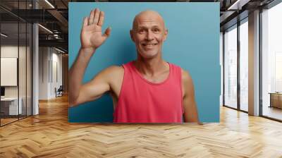 A cheerful bald man in a pink tank top waving at the camera against a blue background, exuding friendliness and positivity. His casual attire and smile add to the inviting demeanor. Wall mural