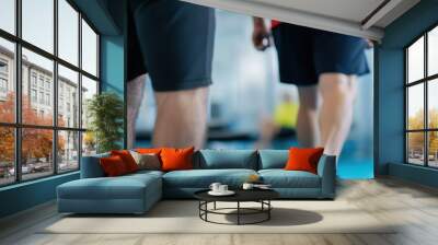 Close-up view focusing on the legs of two individuals walking on a gym floor, suggesting physical activity, fitness routines, and a modern indoor workout environment. Wall mural