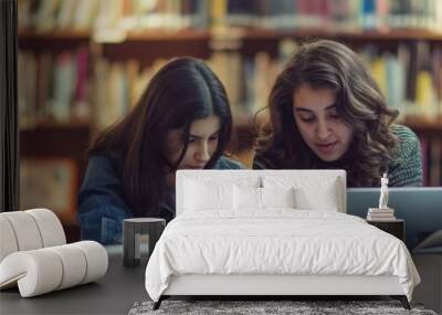 Two focused young women using laptops for studying in a library with shelves filled with books Wall mural