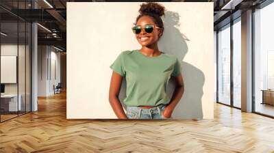 A cheerful woman with sunglasses and a green shirt smiles while leaning against a sunlit wall, enjoying a warm and sunny day outdoors.
 Wall mural