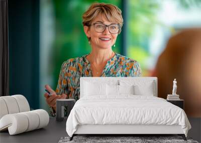 A senior businesswoman with glasses and short hair leads a meeting with enthusiasm, engaging with participants in a modern office.
 Wall mural