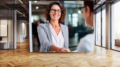 Happy mid aged business woman manager handshaking greeting client in office. Smiling female executive making successful deal with partner shaking hand at work standing at meeting table. Wall mural