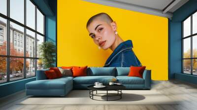 Stylish woman with a shaved head, wearing a denim jacket and flower earrings, posing confidently against a vibrant yellow background. Wall mural