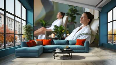 Two women relax in white robes in a spa setting with candles and plants. Wall mural