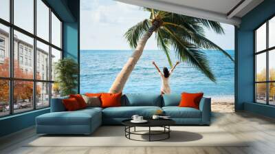 Back view of happy young woman enjoy her tropical beach vacation standing under palm tree Wall mural