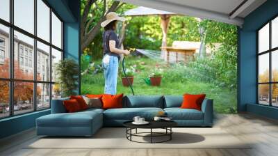 Young woman in hat watering flowers and plants in garden with hose in sunny blooming backyard Wall mural