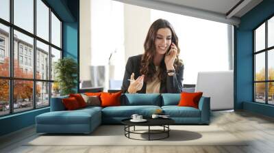 Keep in touch with clients. Shot of a young businesswoman sitting at office desk in front of laptop and making call while onsidering the possibilities with her client. Wall mural