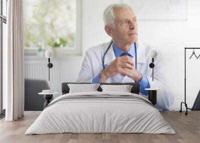 Thinking male doctor sitting in at desk behind computers in the consulting room. Wall mural