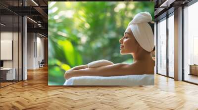 Woman with a towel on her head relaxes on a white towel, looking peaceful and serene with a lush green background. Wall mural