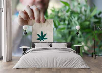 Male hand hold paper packet with marijuana symbol closeup background. Online shopping sativa concept Wall mural