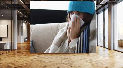 Portrait of of girl sitting in living room covered in blanket and suffering from common cold. Woman having severe flu at home indoors and sneezing into tissue. Hard virus concept Wall mural