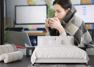 Sick young woman sits in warm blanket with cup at workplace Wall mural