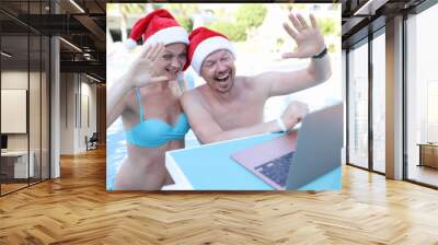 Young man and woman in santa claus hats swimming in pool and waving at laptop screen Wall mural