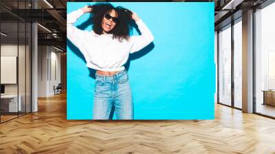 Beautiful black woman with afro curls hairstyle. Smiling model dressed in white summer top and jeans clothes. Sexy carefree female posing near blue wall in studio. Tanned and cheerful. Isolated Wall mural