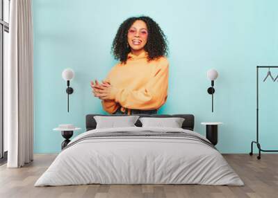 Beautiful black woman with afro curls hairstyle.Smiling model in orange hoodie and trendy jeans clothes. Sexy carefree female posing near blue wall in studio in sunglasses Wall mural