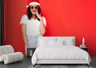 Beautiful woman celebrating New Year.Happy gorgeous female in casual clothes having fun at New Year's Eve party.Holiday celebration.Charming model in Santa Claus near red wall. Shocked and surprised Wall mural