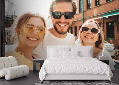 Group of young three stylish friends in the street.Man and two cute girls dressed in casual summer clothes.Smiling models having fun in sunglasses.Women and guy making photo selfie on smartphone Wall mural