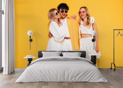 Group of young three stylish friends posing in studio. Fashion man and two cute female dressed in casual summer clothes. Smiling models having fun. Cheerful women and guy near yellow wall, sunglasses Wall mural