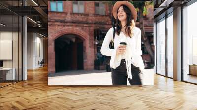 Lifestyle portrait of young stylish woman dressed in white blouse. Model walking with coffee cup in sunny day. Beautiful smiling female in hat. Cheerful and happy. Takeaway coffee Wall mural