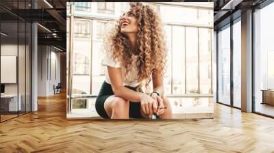 Portrait of beautiful smiling model with afro curls hairstyle dressed in summer hipster clothes.Sexy carefree girl sitting in the street background. Trendy funny and positive woman having fun Wall mural