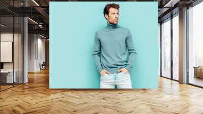 Portrait of handsome confident  model. Sexy stylish man dressed in  sweater and jeans. Fashion hipster male with curly hairstyle posing near blue wall in studio. Isolated Wall mural