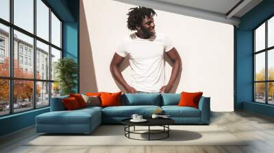 Portrait of handsome hipster model.Unshaven African man dressed in white summer t-shirt and jeans. Fashion male with dreadlocks hairstyle posing near white wall in the street Wall mural