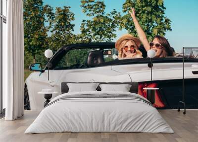 Portrait of two young beautiful and smiling hipster female in convertible car. Sexy carefree women driving. Positive models riding and having fun outdoors. Enjoying summer days. Cheerful and happy Wall mural