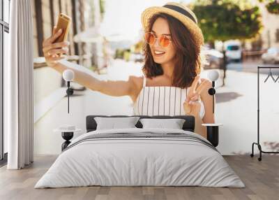 Teen smiling girl in summer white hipster clothes in taking selfie on smartphone on warm sunset background. Showing peace sign Wall mural