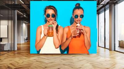 Two beautiful sexy smiling hipster women in summer colorful swimwear bathing suits.Trendy girls in sunglasses.Going crazy.Funny models isolated on blue.Drinking fresh cocktail smoozy drink Wall mural