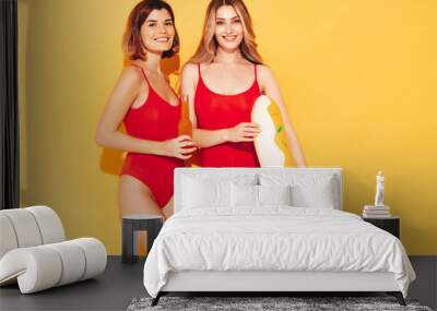 Two beautiful sexy smiling hipster women in summer swimwear bathing suits. Trendy models in studio. Hot female isolated on yellow. In sunglasses, holding bottle of lemonade, donut inflatable mattress Wall mural