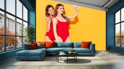 Two beautiful sexy smiling hipster women in summer swimwear bathing suits. Trendy models in studio. Hot female isolated on yellow. In sunglasses, holding smartphone. Taking selfie photos Wall mural