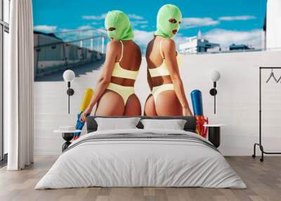 Two beautiful sexy women in green underwear. Models wearing bandit balaclava mask. Hot seductive female in nice lingerie posing in the street at sunny day, blue sky. Crime and violence. Hold water gun Wall mural